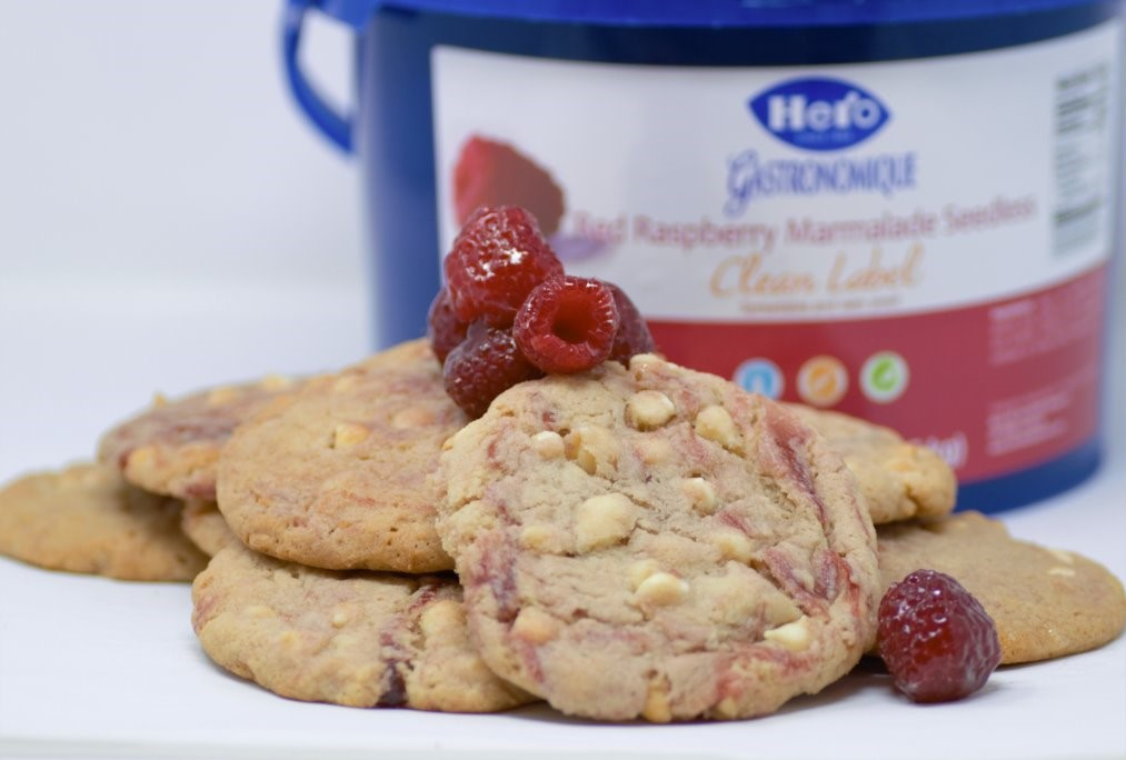 Cookies with white chocolate chips and raspberries on top. The cookies are placed in front of the Hero Raspberry Bakery Jam.